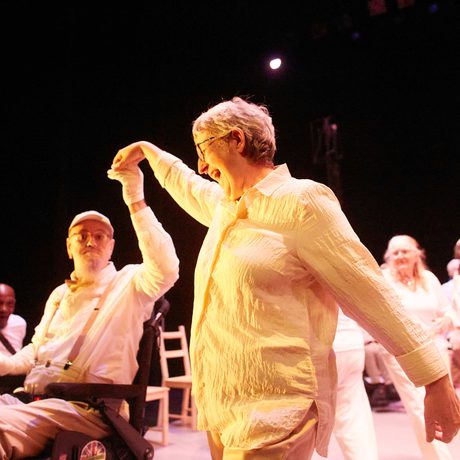 Two older performers dressed in white dance together, one White woman with grey hair and one White man in a wheelchair with a grey beard.