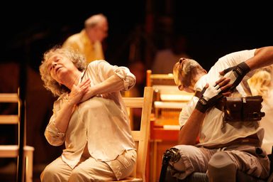 Two older performers lean back with hands over their hearts, one white woman on a wooden chair and one black man in a wheelchair.