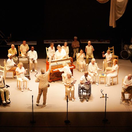 A stage of older performers in white sitting on wooden chairs or in wheelchairs. They play various instruments.