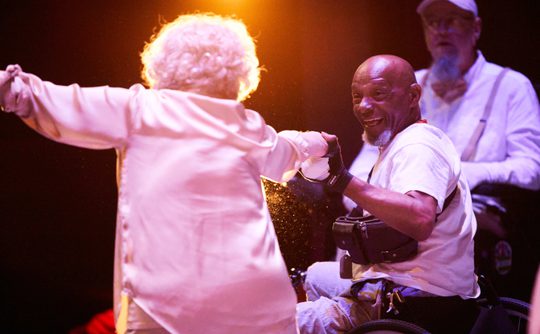 Two older performers dressed in white dance together, one White woman with grey hair and one bald Black man in a wheelchair.