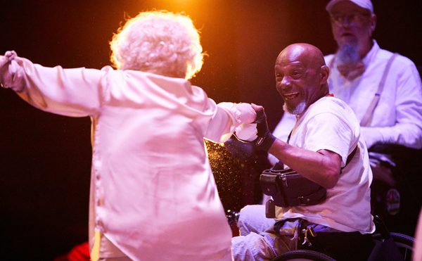 Two older performers dressed in white dance together, one White woman with grey hair and one bald Black man in a wheelchair.