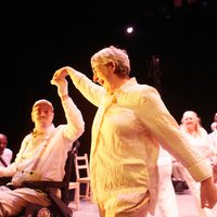 Two older performers dressed in white dance together, one White woman with grey hair and one White man in a wheelchair with a grey beard.