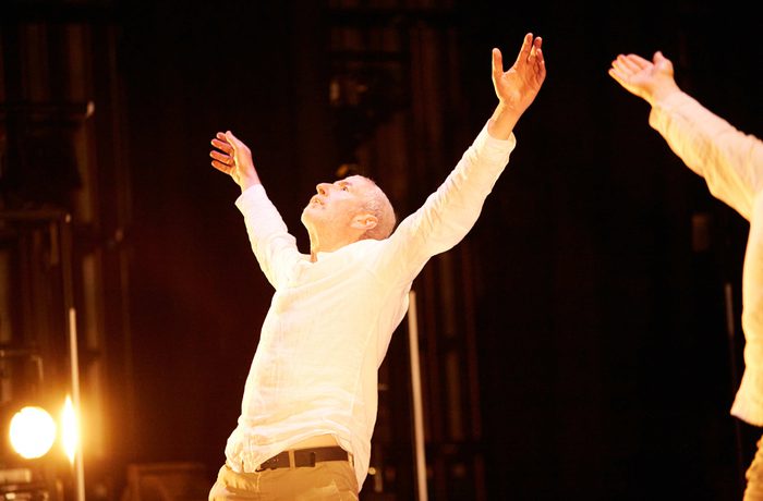 An older White male performer wearing white with his arms raised and head tilted back.