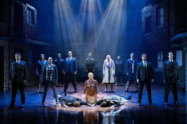 A stage of performers in dark clothing standing behind a woman kneeling in the spotlight between two seemingly-dead young men.