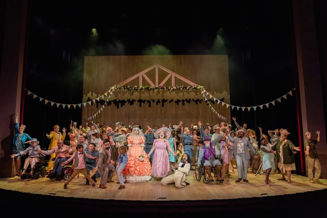 Large cast on stage with arms outstretched mid song barring one character in a peach wedding dress who looks dismayed. The cast is made up of local people from a variety of age ranges.