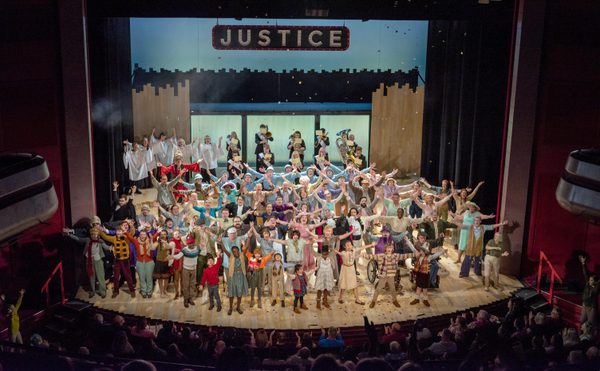 A lively theater stage filled with a diverse cast in colorful costumes, under a "Justice" sign, arms outstretched joyfully, conveying celebration. Performance of The Doncastrian Chalk Circle 2022.