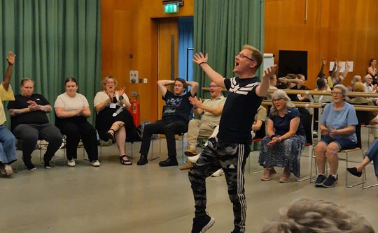 A man stands in a rehearsal room surrounded by seated people. He has his arms spread in a lively performance as everyone around him cheers.