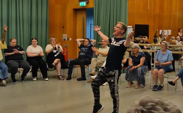 A man stands in a rehearsal room surrounded by seated people. He has his arms spread in a lively performance as everyone around him cheers.