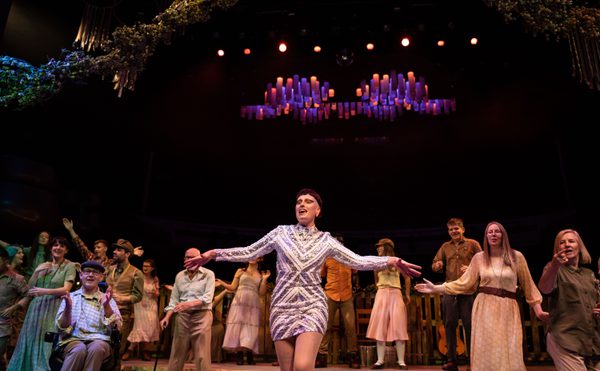 A performer in a sequined dress stands centre stage with arms outstretched, surrounded by a group of people in earthy-toned costumes under warm stage lighting.