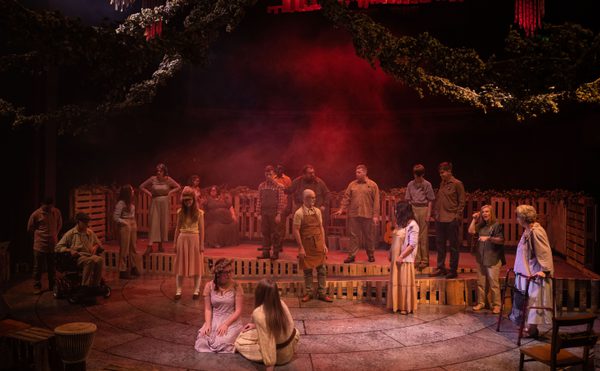 Theatrical scene with diverse performers in rustic costumes on stage surrounded by wooden crates, under dim red lighting and greenery, creating a tense, dramatic atmosphere.