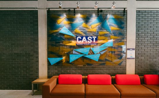 A brick wall with a plastic board hanging from it decorated with blue and yellow triangles and the Cast logo.