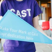 A person in a purple t-shirt with a Cast logo holds a blue triangle with the words "Make your mark at Cast, your dedication here" on it.