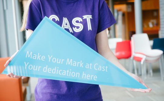 A person in a purple t-shirt with a Cast logo holds a blue triangle with the words "Make your mark at Cast, your dedication here" on it.