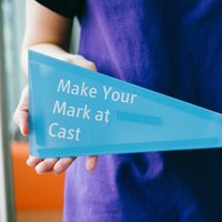 A person in a purple t-shirt holds a blue triangle with the words "Make your mark at Cast" on it.