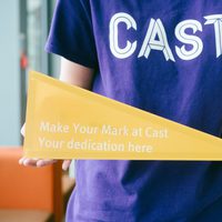 A person in a purple t-shirt with a Cast logo holds a yellow triangle with the words "Make your mark at Cast, your dedication here" on it.