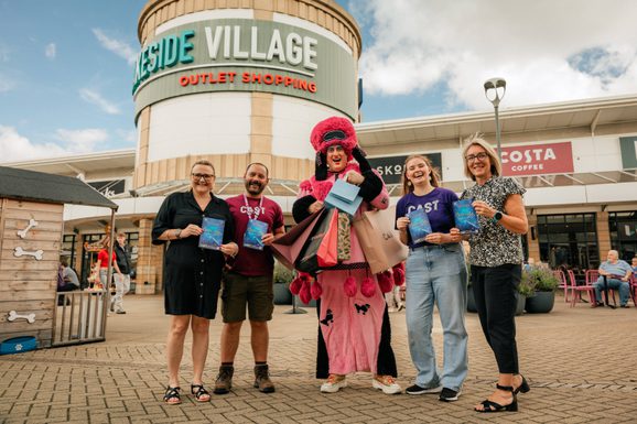 A group of five people, one in a colorful costume, joyfully pose holding bright flyers that say "Cinderella" in front of Lakeside Village Outlet Shopping. Clear sky above.