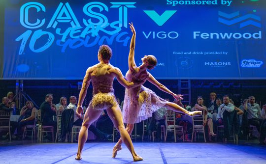 Two ballet dancers holding a pose in front of an audience sitting at formal dinner tables on a theatre stage.