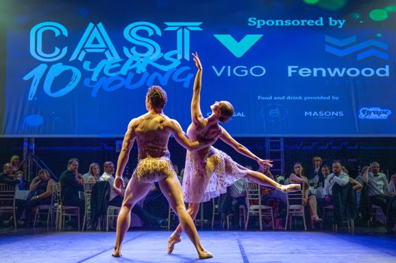 Two ballet dancers holding a pose in front of an audience sitting at formal dinner tables on a theatre stage.