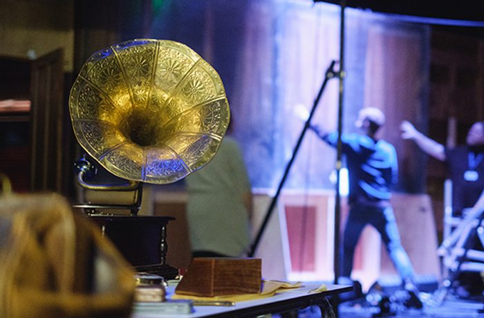 Gramophone sits to the front of the photo on a table, behind two blurred figures put up stage set.