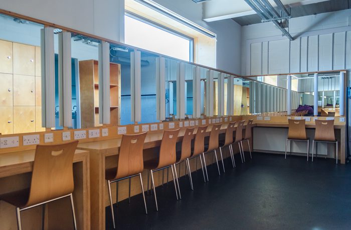 A brightly-lit dressing room with a long wooden table, ten sleek wooden chairs, and a large mirror. Power outlets line the table, creating a studio-like ambiance.