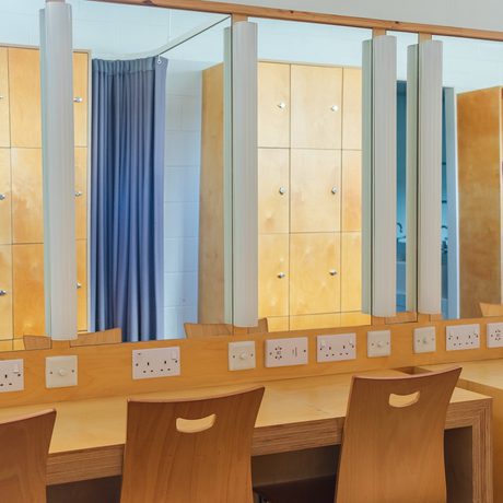 A brightly lit dressing room with wooden chairs and tables facing large mirrors. The walls feature wooden cabinets, and power outlets line the table.