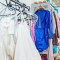 Racks of colorful costumes and bridal gowns hang in a backstage dressing room. The vibrant blue sequined outfit stands out among the whites and pastels.
