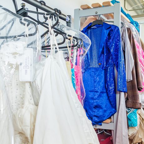 Racks of colorful costumes and bridal gowns hang in a backstage dressing room. The vibrant blue sequined outfit stands out among the whites and pastels.