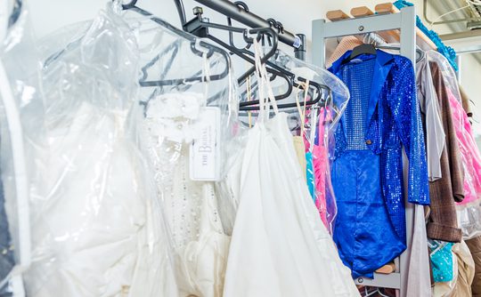Racks of colorful costumes and bridal gowns hang in a backstage dressing room. The vibrant blue sequined outfit stands out among the whites and pastels.