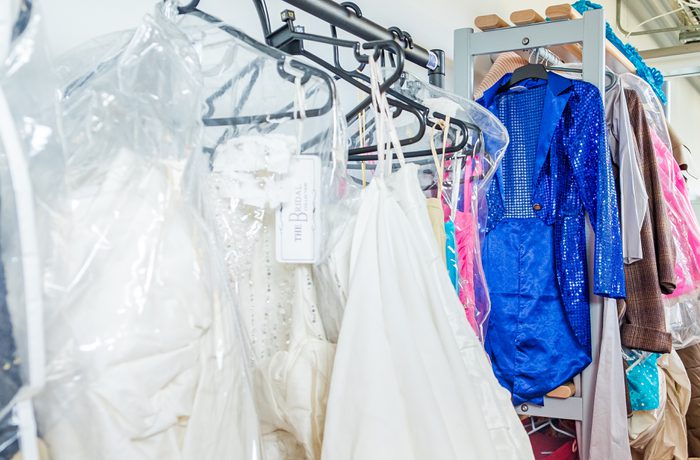 Racks of colorful costumes and bridal gowns hang in a backstage dressing room. The vibrant blue sequined outfit stands out among the whites and pastels.