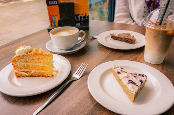 A table with three dessert plates: a slice of layered orange cake, a berry-topped cheesecake, and a chocolate pastry. Nearby are a cappuccino and an iced coffee. The mood is cozy and inviting.