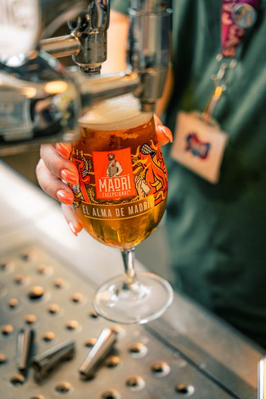 A hand with bright coral nails holds a glass of Madri beer under a tap. The vibrant glass design reads "El Alma de Madrid," exuding a lively vibe.