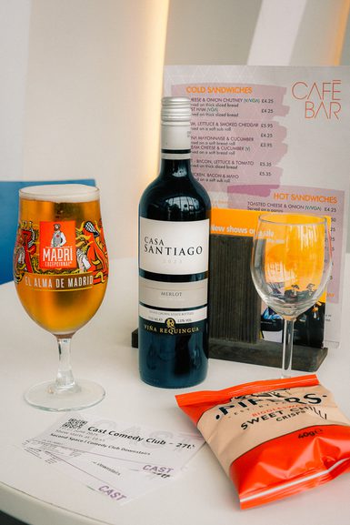 A cafe table displays a wine bottle, full beer glass, empty wine glass, sandwich menu, comedy club tickets, and a snack bag, creating a relaxed vibe.
