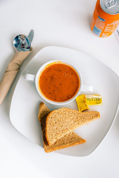 A white plate with a cup of tomato soup, two slices of toasted bread, and a butter packet. A rolled napkin, spoon, and orange Fanta can are nearby.