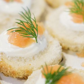 Round canapés with creamy white spread, topped with salmon pieces and fresh dill. Served on a wooden board, they convey an elegant, appetizing tone.