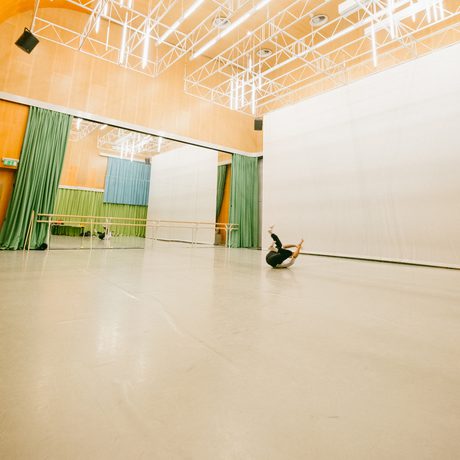 The spacious Dance Space at Cast with high ceilings and bright lights. A dancer lies on the floor mid-move, surrounded by mirrors and green curtains. The mood is focused and artistic.