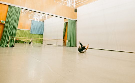 The spacious Dance Space at Cast with high ceilings and bright lights. A dancer lies on the floor mid-move, surrounded by mirrors and green curtains. The mood is focused and artistic.