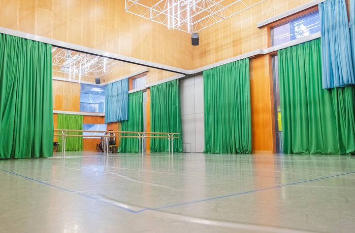 Spacious dance studio with green and blue curtains, mirrored walls, and wooden paneling. A ballet barre lines the wall, creating a calm, focused ambiance.
