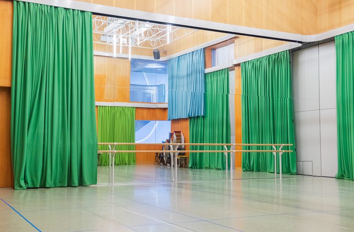 A spacious dance studio with green curtains, mirrored walls, and a barre. Stacked chairs in the corner. The tone is bright and inviting.