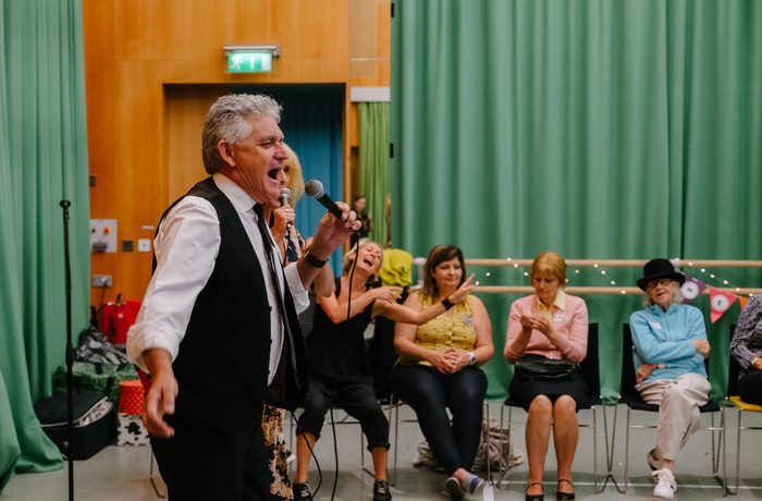 A man passionately sings into a microphone in a room with green curtains. Seated audience members, smiling and clapping, suggest a lively atmosphere during a Sing, Swing and Cake group at Cast in Dance Space..