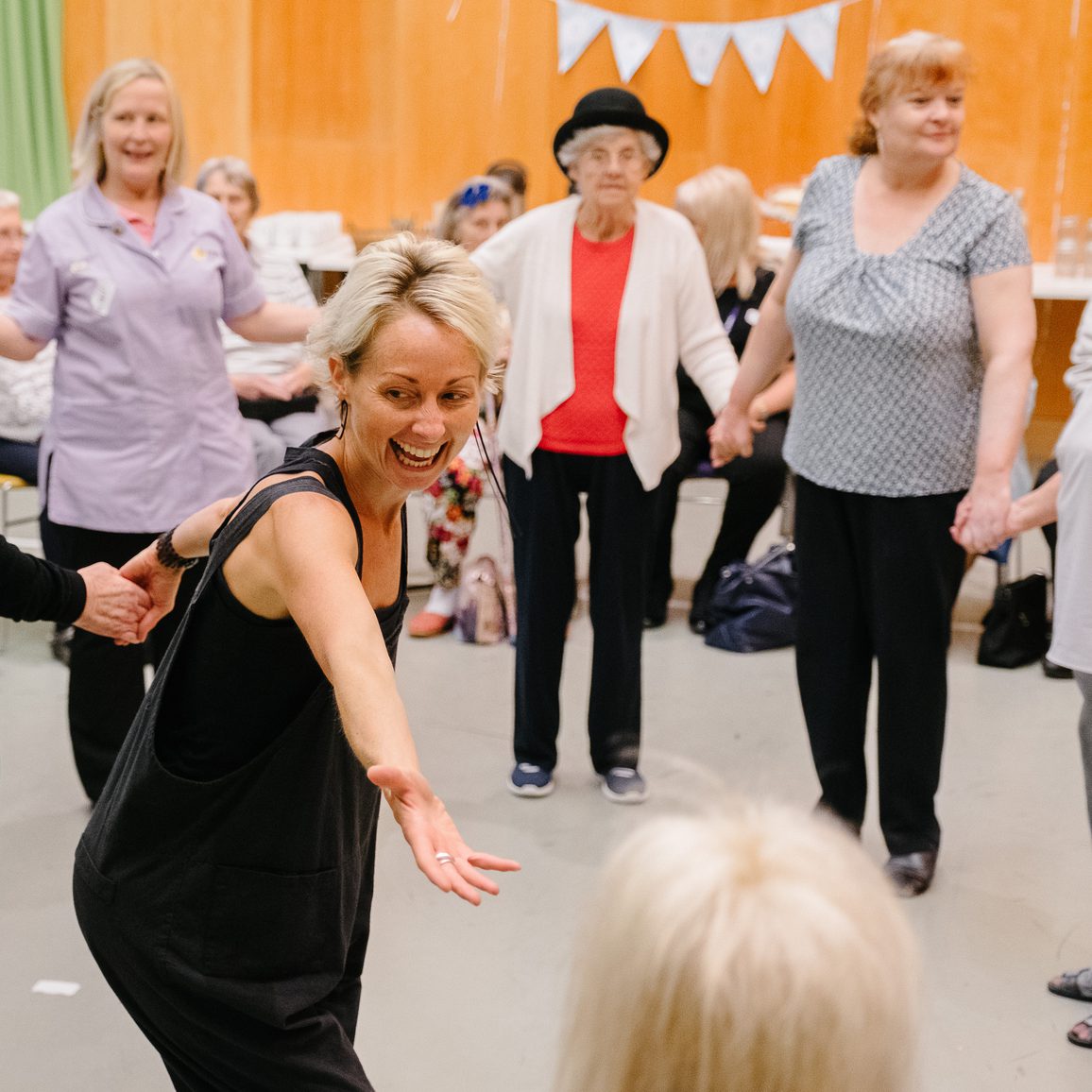 A joyful group dance during Cast's Sing, Swing and Cake group. A smiling woman in the foreground reaching out to someone just off camera, older people holding hands in a circle, vibrant energy, and spring decor in Dance Space.