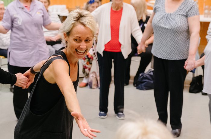 A joyful group dance during Cast's Sing, Swing and Cake group. A smiling woman in the foreground reaching out to someone just off camera, older people holding hands in a circle, vibrant energy, and spring decor in Dance Space.