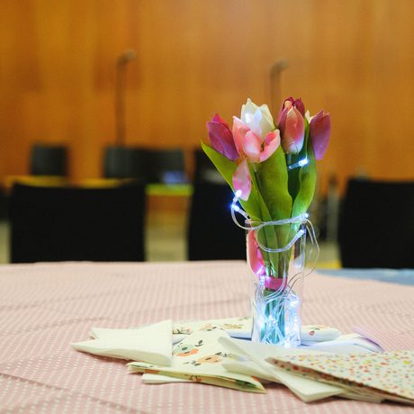 A vase with pink tulips wrapped in fairy lights is centered on a patterned tablecloth, surrounded by folded napkins. Chairs are blurred in the background of Cast's Dance Space.