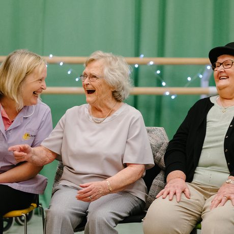 Three women seated and laughing together in a cheerful atmosphere. One wears a carer's uniform. Green background with faint string lights, conveying joy during a session of Sing, Swing and Cake in Cast's Dance Space.
