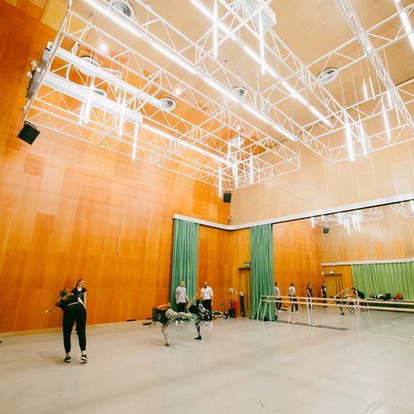 A spacious dance studio with high ceilings and mirrored walls. Several people practice dance moves, surrounded by green curtains and warm wood panels in Cast's Dance Space.