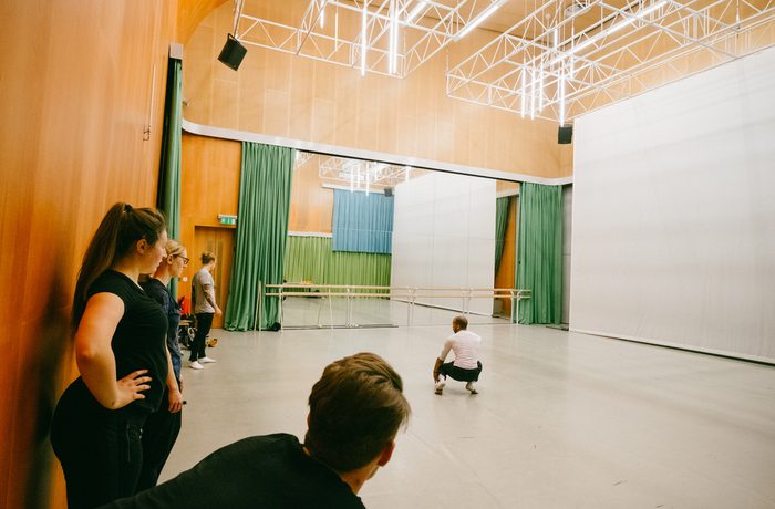 A spacious dance studio with wooden walls and green curtains. Several people observe as one person squats on the floor, near large mirrors and ballet bars in Cast's Dance Space.