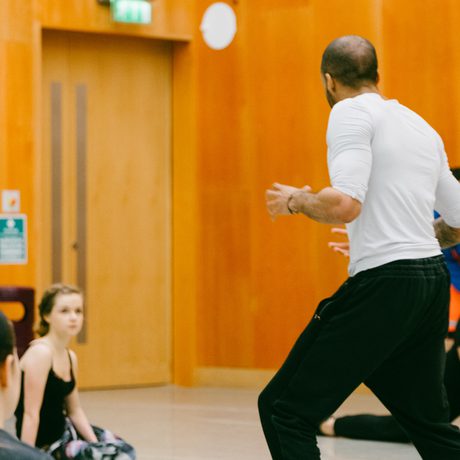 A dance instructor in a white shirt and black pants leads a class of seated students in Cast's Dance Space with wooden walls and green curtains, conveying focus and engagement.