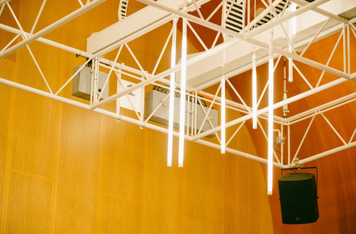 White ceiling beams with fluorescent lights hang in a room with warm wooden paneling. A black speaker is suspended, creating an industrial yet cozy ambiance in Cast's Dance Space.