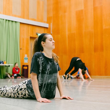 A woman practices yoga in a studio with wood-paneled walls and green curtains. Other participants stretch in the background, creating a calm atmosphere in Cast's Dance Space.