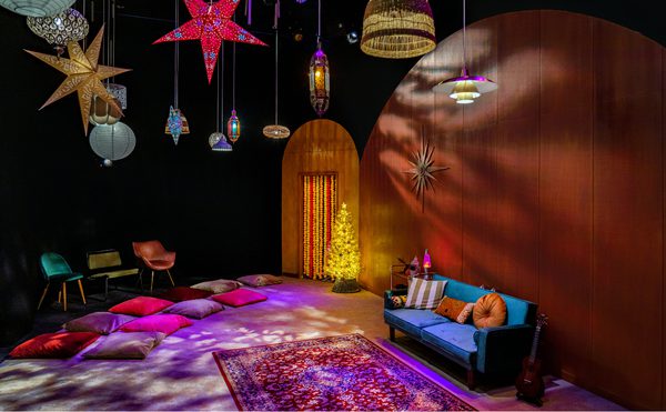 A cozy room with a festive vibe, featuring colorful star lanterns, a glowing tree, cushions on the floor, a blue sofa, and a guitar against warm wood walls. This photo is of the set from cast's 2021 production of Wish, a early years production in Drama Space.