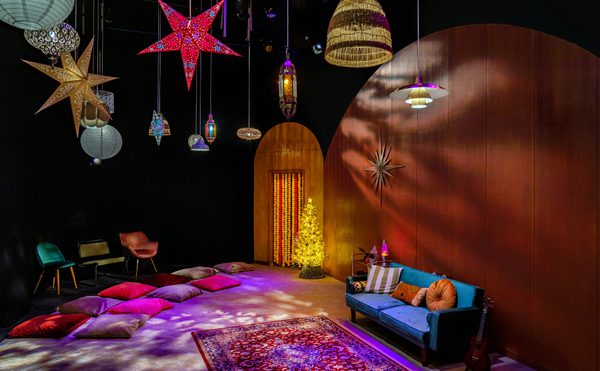 A cozy room with a festive vibe, featuring colorful star lanterns, a glowing tree, cushions on the floor, a blue sofa, and a guitar against warm wood walls. This photo is of the set from cast's 2021 production of Wish, a early years production in Drama Space.